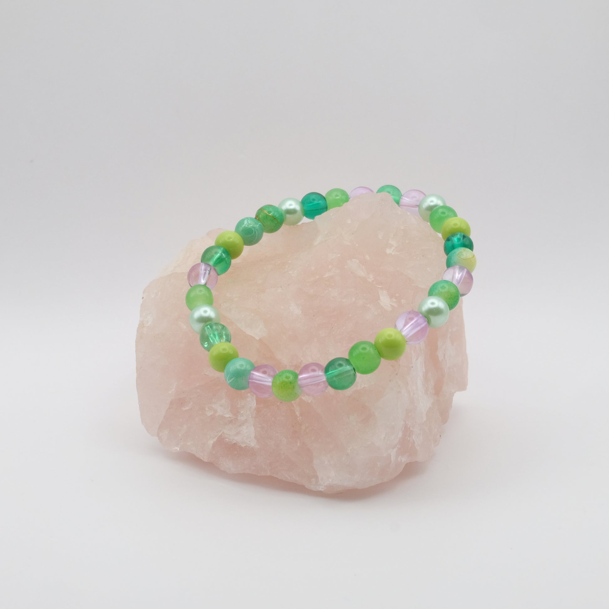 A purple, green and teal elastic beaded bracelet sitting on a rose quartz rock with a white background