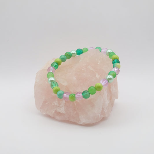 A purple, green and teal elastic beaded bracelet sitting on a rose quartz rock with a white background