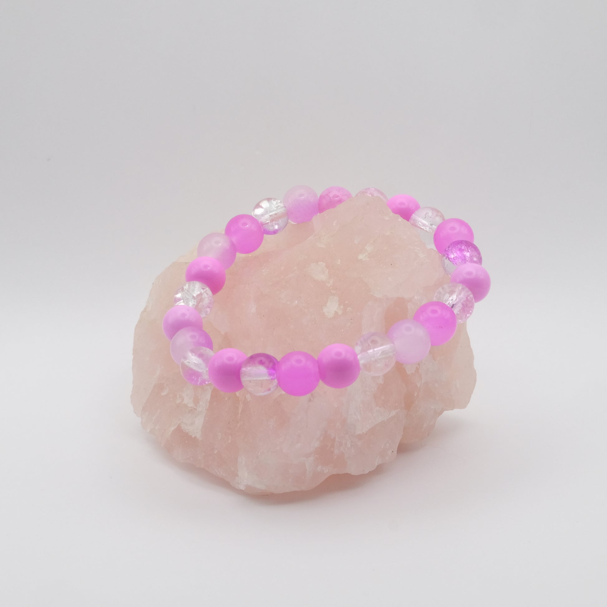 A pinky purple elastic beaded bracelet sitting on a rose quartz rock with a white background