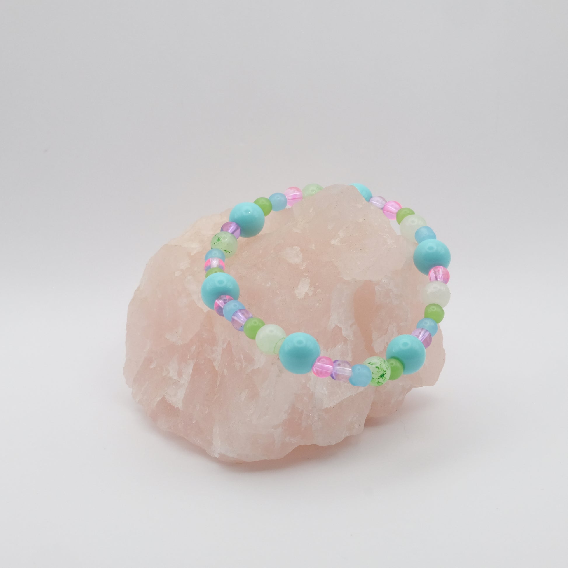 A single elastic beaded bracelet in pink, blue, purple and green sitting on a rose quartz rock with a white background