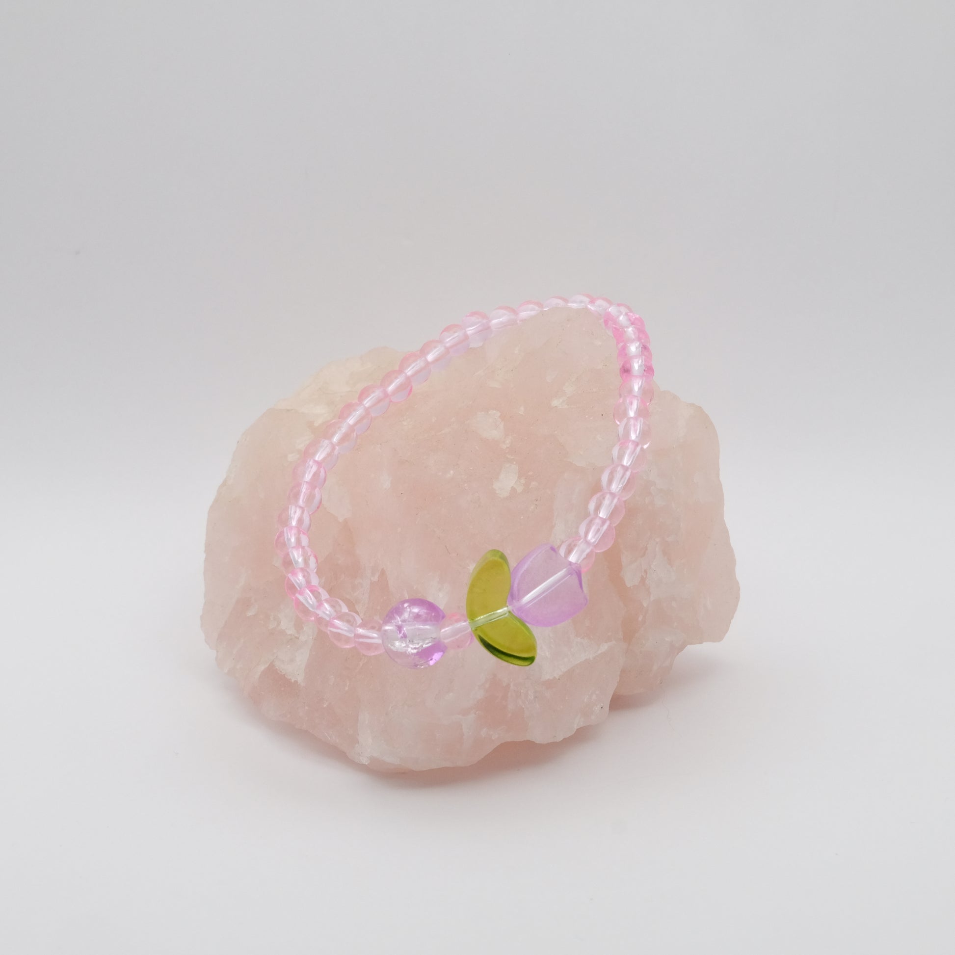 A single elastic beaded bracelet in pink, with a single purple feature bead and a single purple glass flower bead sitting on a rose quartz rock with a white background