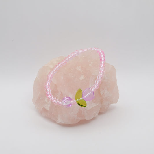 A single elastic beaded bracelet in pink, with a single purple feature bead and a single purple glass flower bead sitting on a rose quartz rock with a white background