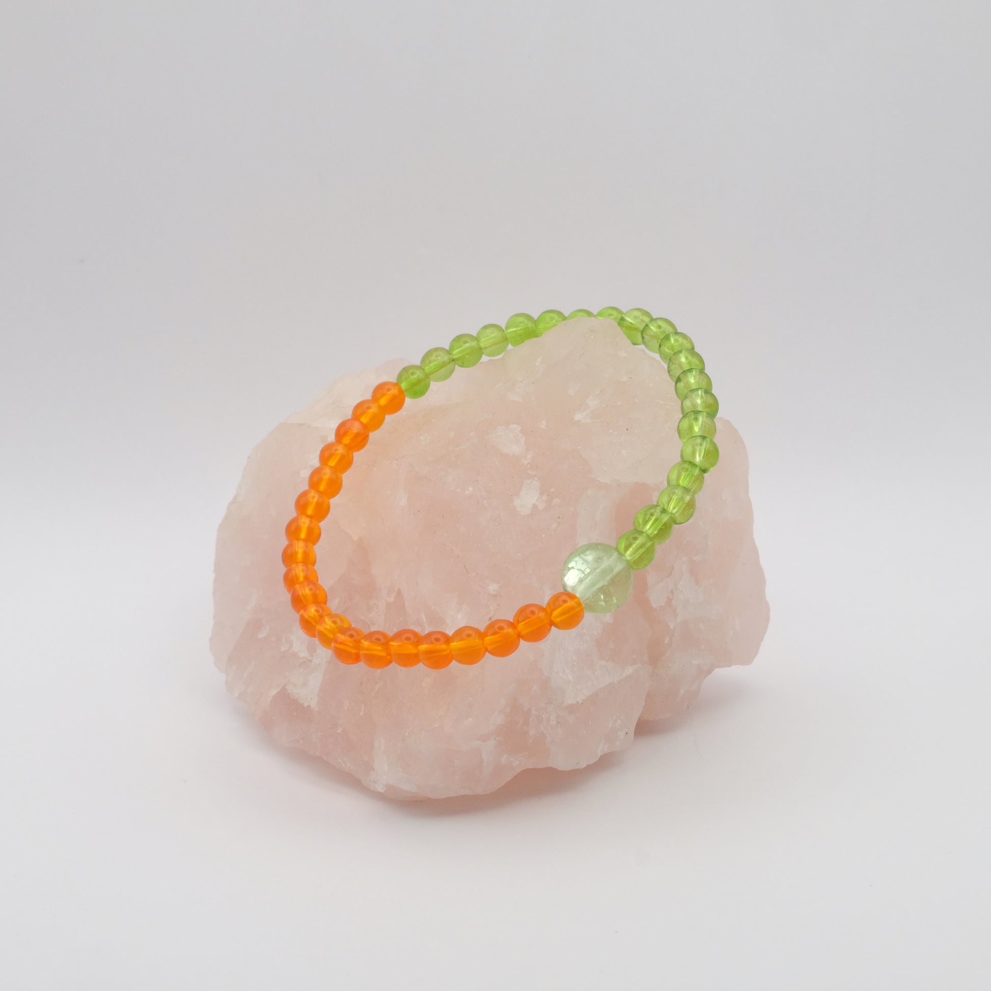 A single elastic beaded bracelet in orange and green sitting on a rose quartz rock with a white background.