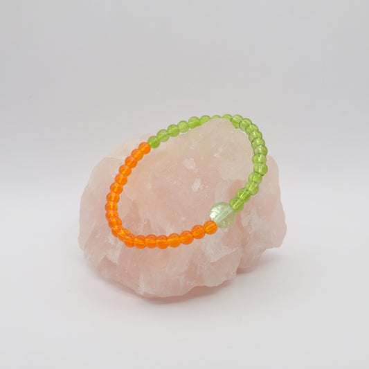 A single elastic beaded bracelet in orange and green sitting on a rose quartz rock with a white background.