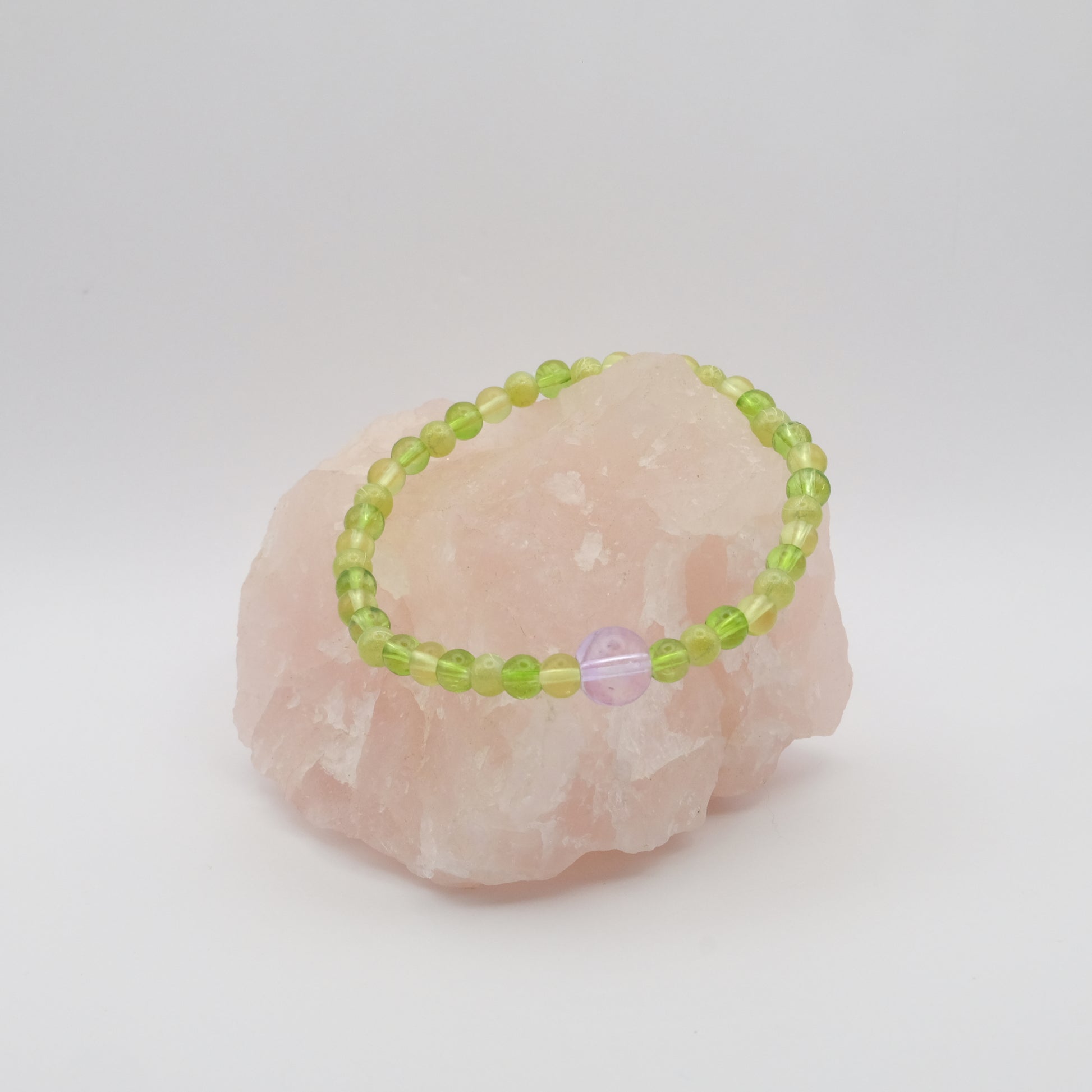 A single elastic beaded bracelet in purple and green sitting on a rose quartz rock with a white background