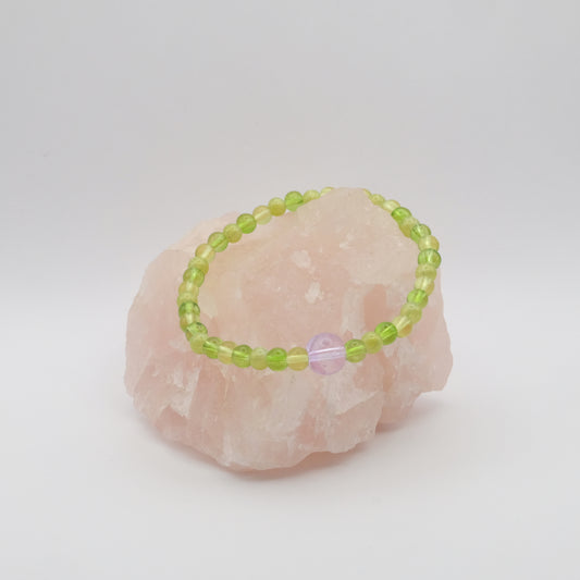 A single elastic beaded bracelet in purple and green sitting on a rose quartz rock with a white background