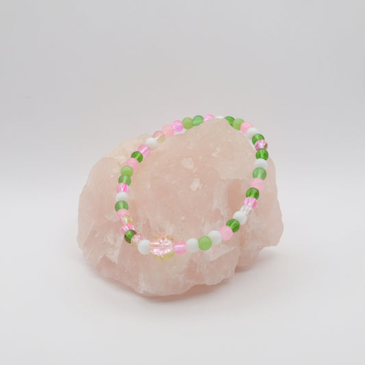 A single elastic beaded bracelet in pink, green and white sitting on a rose quartz rock with a white background