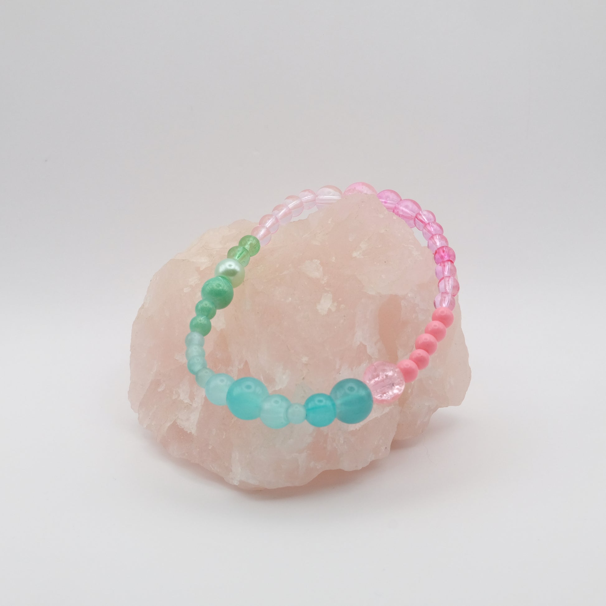 A single elastic beaded bracelet in pink, green and aqua sitting on a rose quartz rock with a white background
