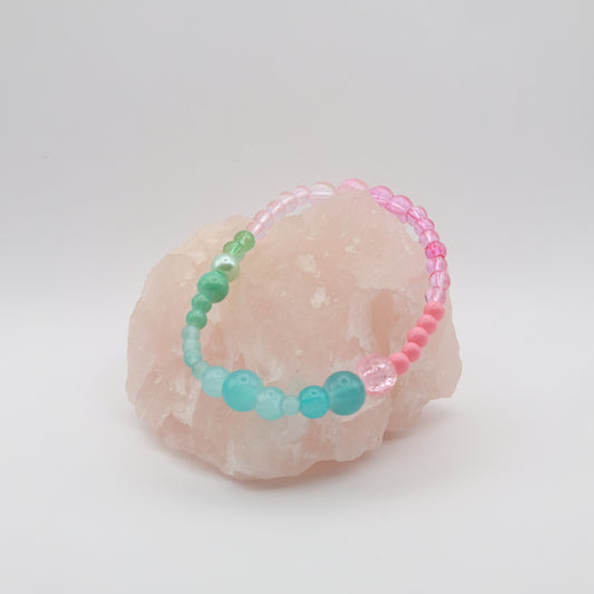 A single elastic beaded bracelet in pink, green and aqua sitting on a rose quartz rock with a white background