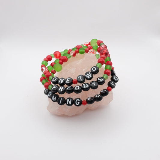 Three red and green beaded bracelets with black letter beads spelling "ONE TWO FREDDYS COMING FOR YOU" on a white background