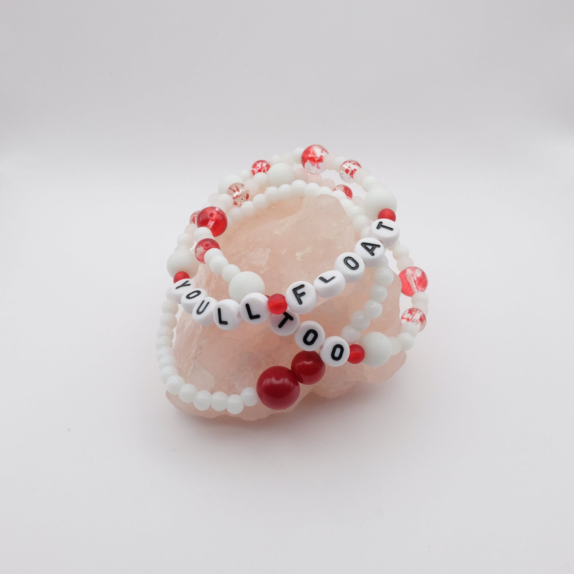 Beaded bracelets with red and white beads on a pink stone with 'YOU'LL FLOAT TOO' text.