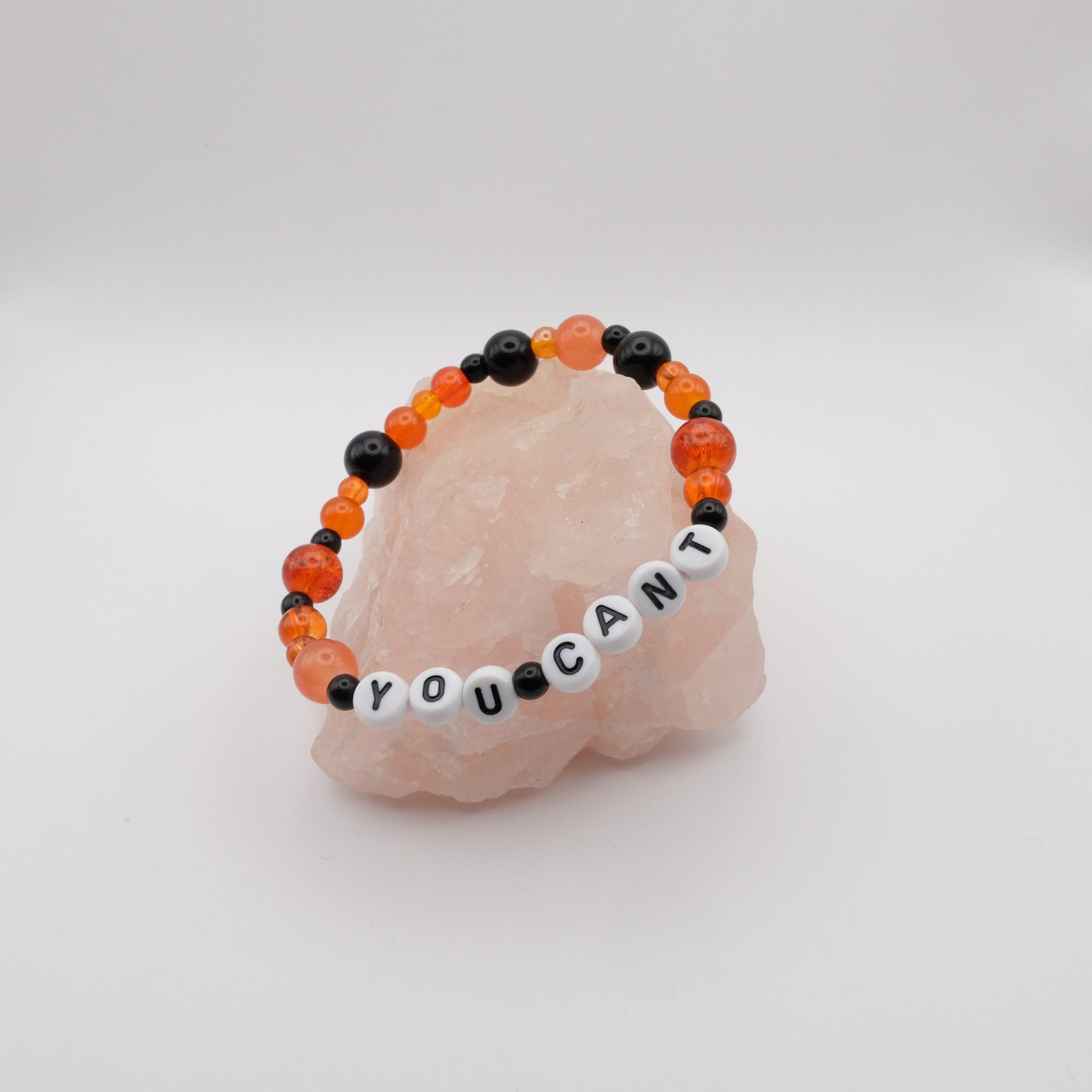 Black  and  orange  bracelet with YOU CANT spelled out in white letter beads, placed on a pink stone