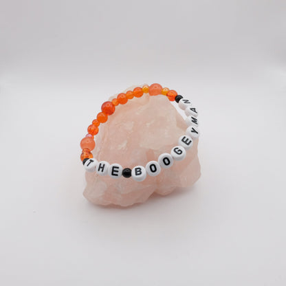 Black and orange bracelet with letter beads spelling THE BOOGEYMAN sitting on a rose quartz rock against a white background