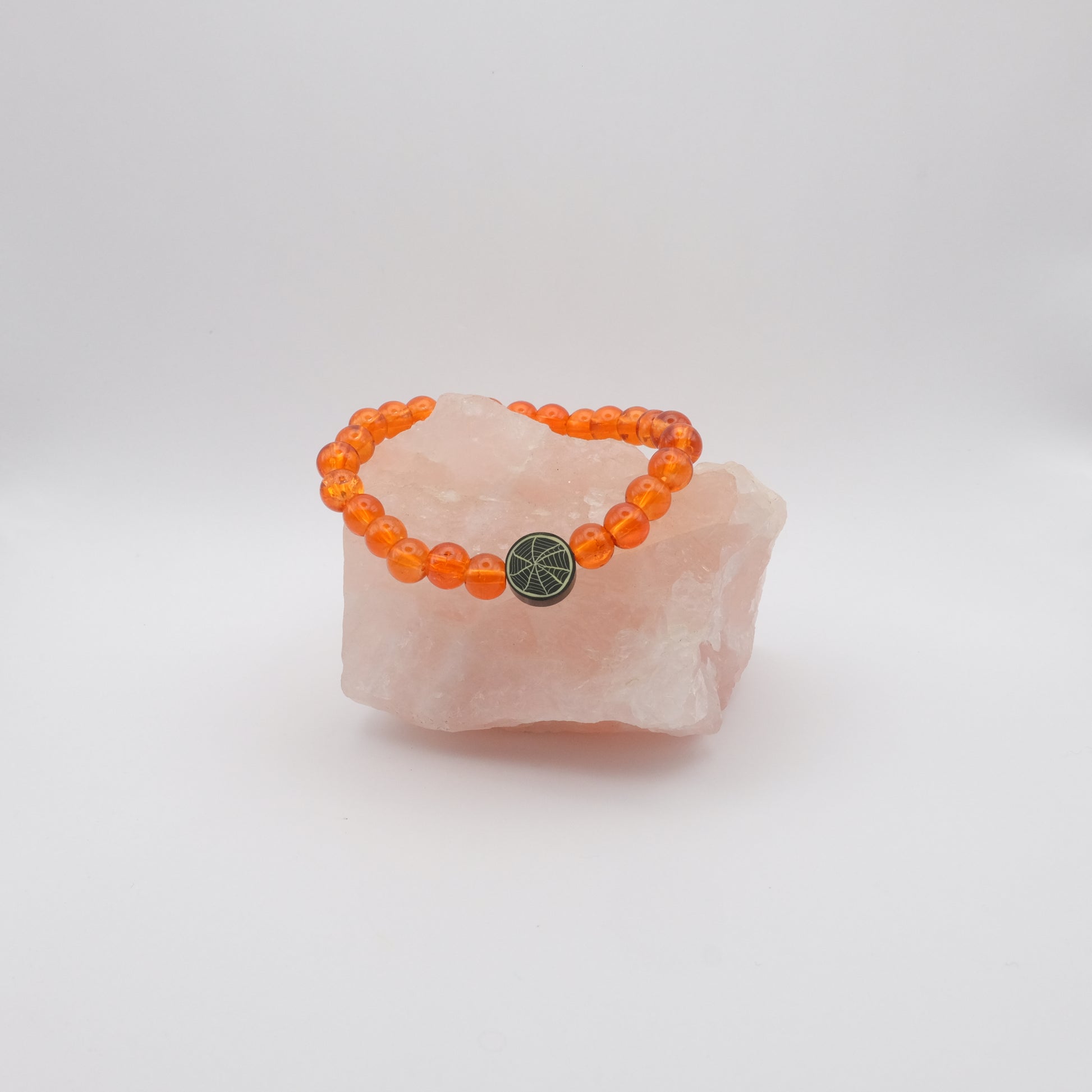 Orange beaded bracelet with a black  spiderweb bead on a pink stone against a white background