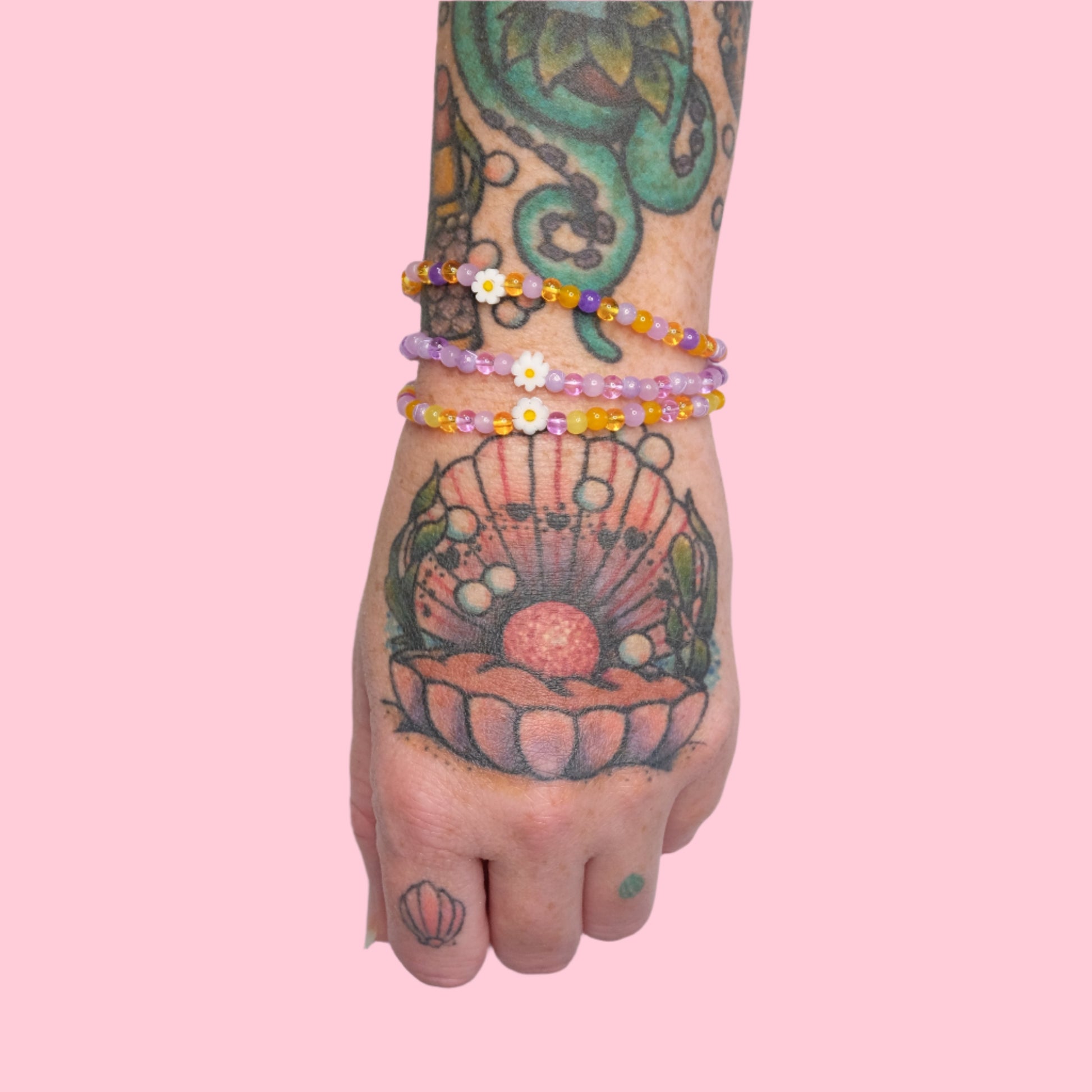 A tattooed arm with octopus and seashell artwork wears three delicate, elastic beaded bracelets in purple and yellow hues, each featuring small white daisy accents, set against a solid pink background.