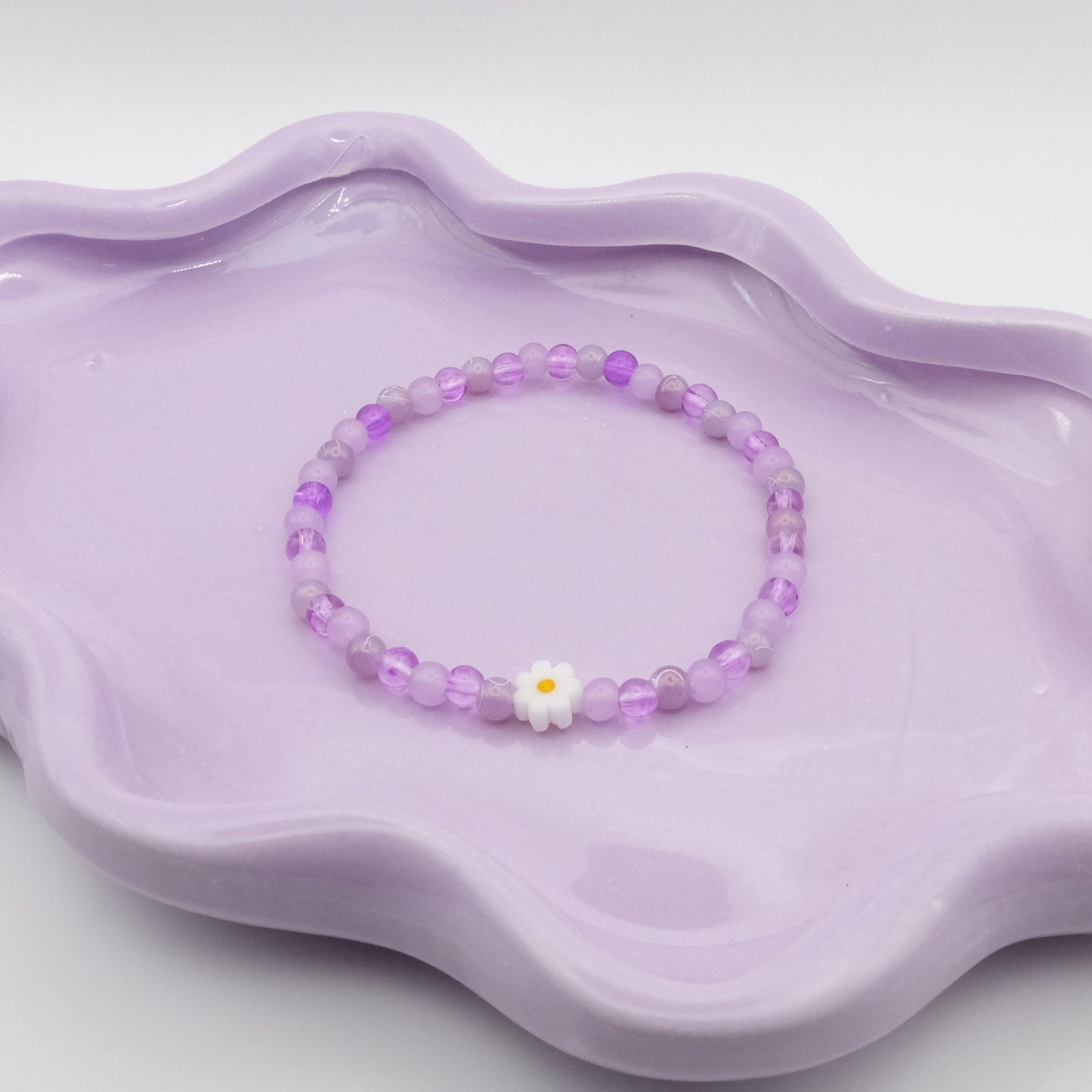 An elastic beaded bracelet crafted entirely from 4mm purple glass beads, finished with a single white daisy bead at the center. It’s displayed on a wavy lavender dish against a simple, light backdrop.
