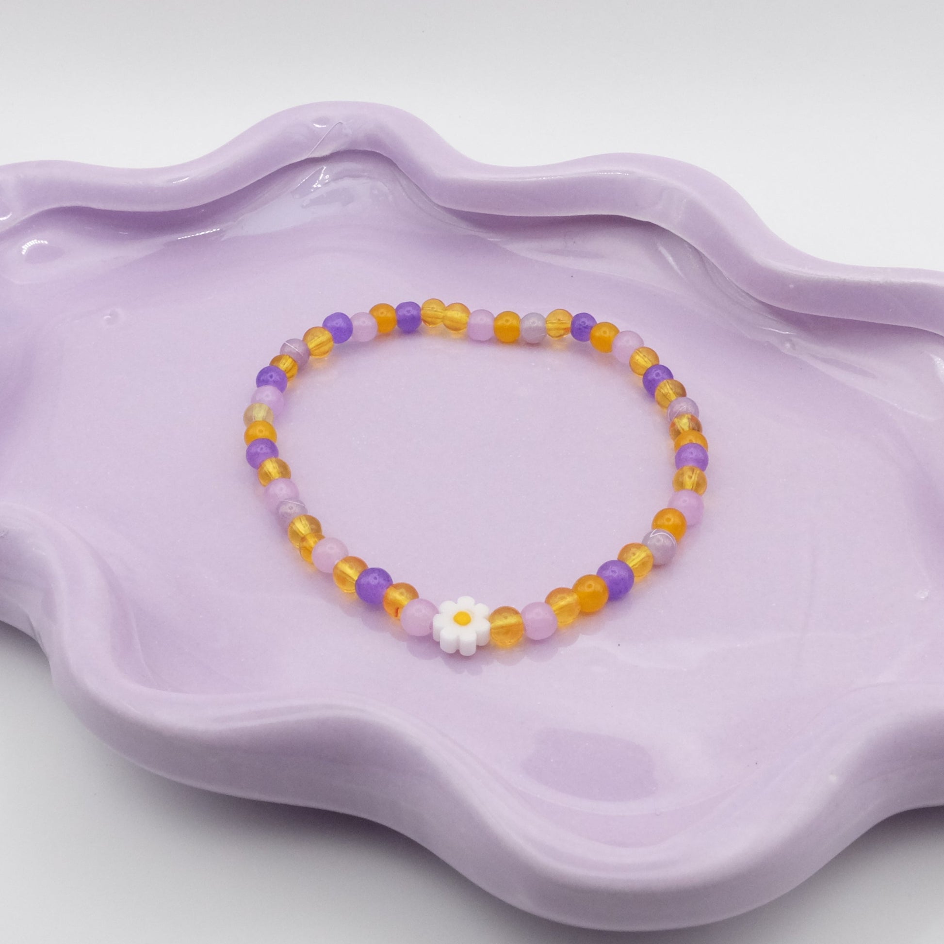 An elastic beaded bracelet featuring 4mm glass beads in a cheerful swirl of light and dark purples plus yellows, accented by a single white daisy bead. Displayed on a wavy lavender dish, it’s set against a minimal, neutral background.