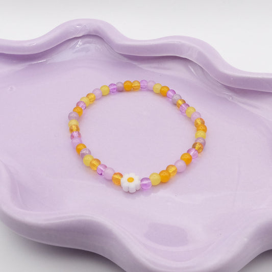 An elastic beaded bracelet made from 4mm glass beads in a glowing mix of yellows and purples, highlighted by a single white daisy bead. Photographed on a wavy lavender dish, it’s shown against a clean, light background