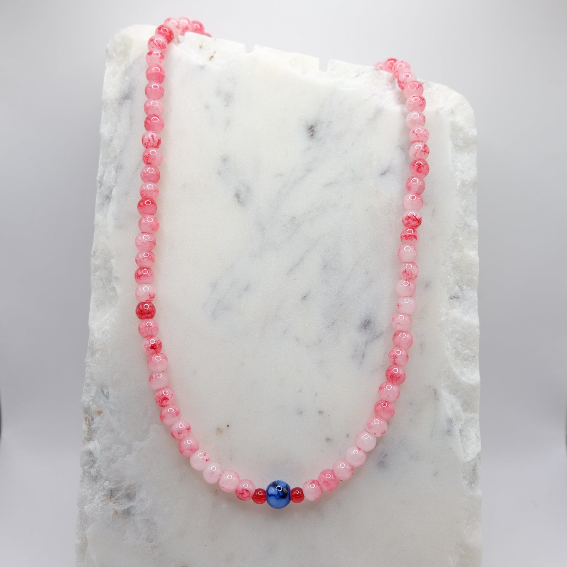 A beaded necklace composed of 6mm red and white marbled glass beads - featuring an 8mm blue and white marbled centre bead with small red accents, shown on a white marble background.