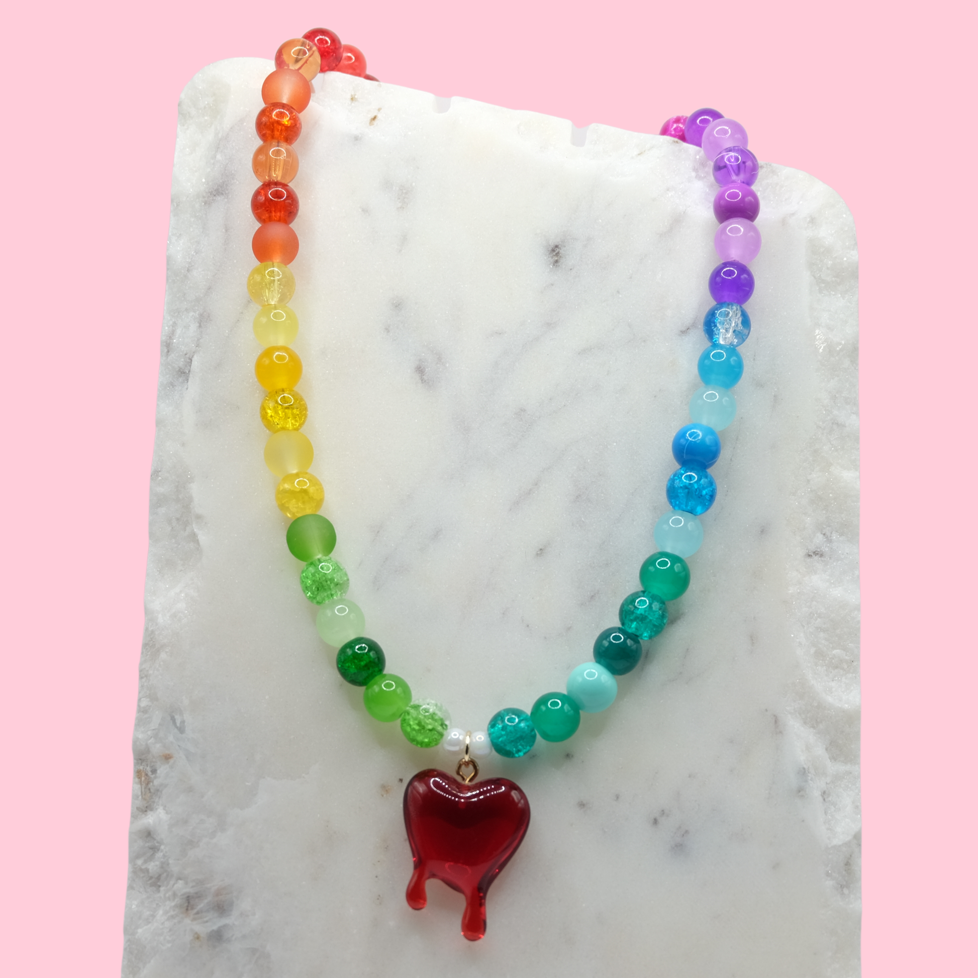 A handmade beaded necklace composed of rainbow-toned 8mm glass beads in mixed glossy, frosted, and crackle finishes, featuring a red resin bleeding heart charm at the centre, displayed on a white marble slab.