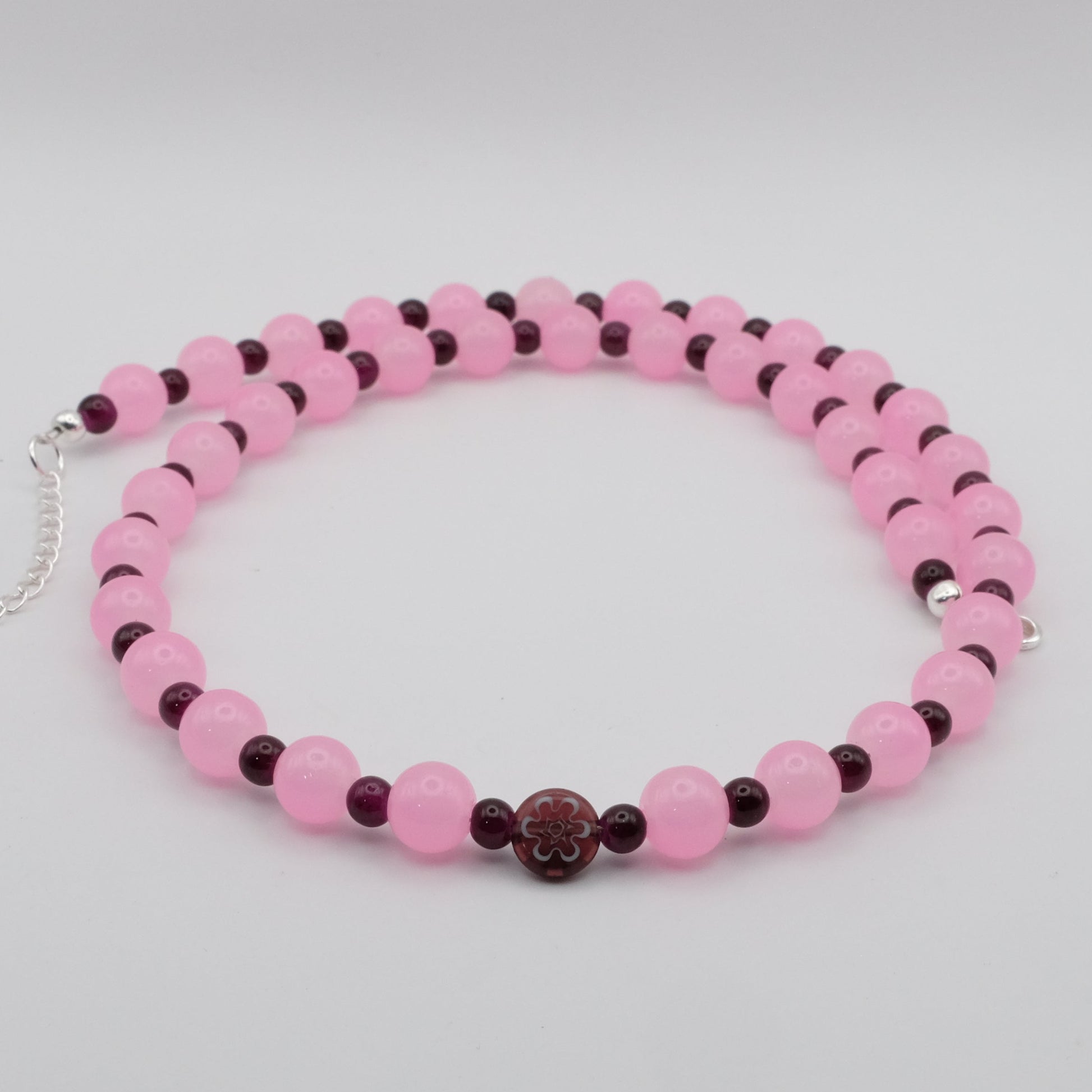 A handmade beaded necklace featuring alternating 8mm pink glass beads and 4mm deep plum glass beads with a flat glass flower focal bead, displayed on a white background.