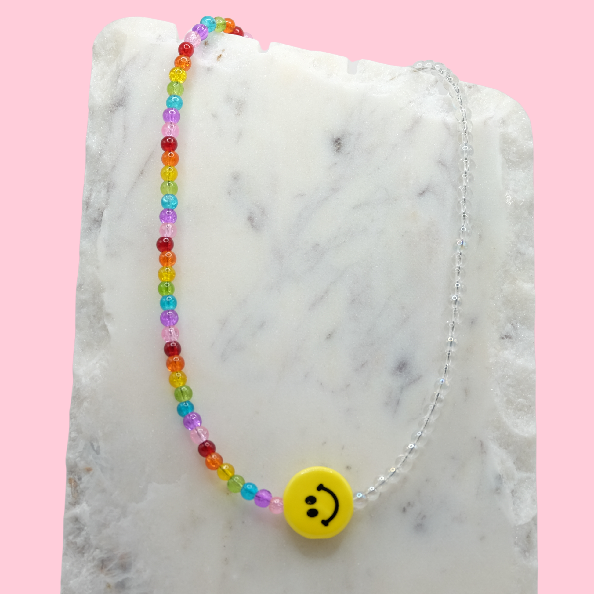 A handmade beaded necklace composed of clear 4mm glass beads and rainbow-toned 4mm glass beads, featuring a yellow ceramic smiley face bead at the centre, displayed on a white marble slab.