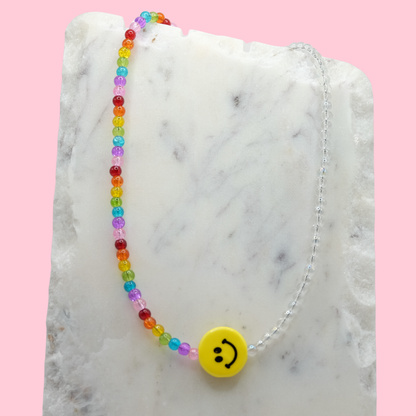 A handmade beaded necklace composed of clear 4mm glass beads and rainbow-toned 4mm glass beads, featuring a yellow ceramic smiley face bead at the centre, displayed on a white marble slab.