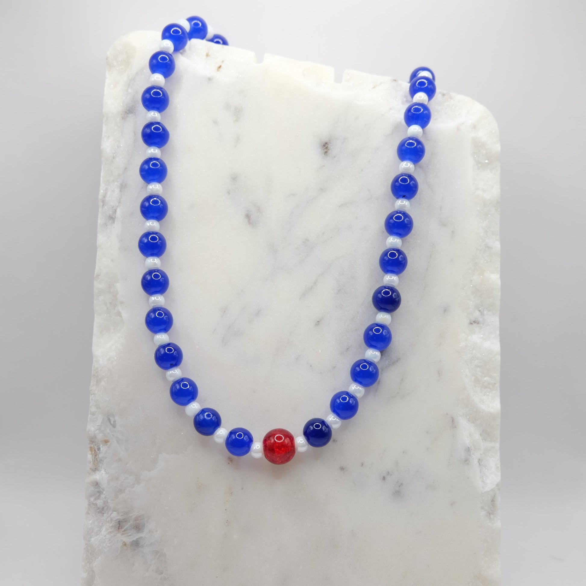 A beaded necklace composed of blue glass beads and metallic white beads - featuring a central red crackle glass bead, shown on a white marble background.