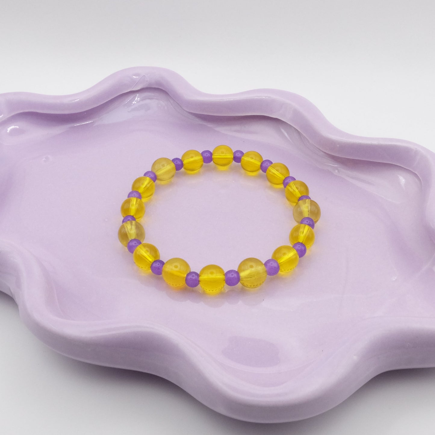 An elastic beaded bracelet featuring alternating 8mm transparent yellow glass beads and 6mm dark purple glass beads, displayed on a wavy lavender ceramic dish against a light backdrop.