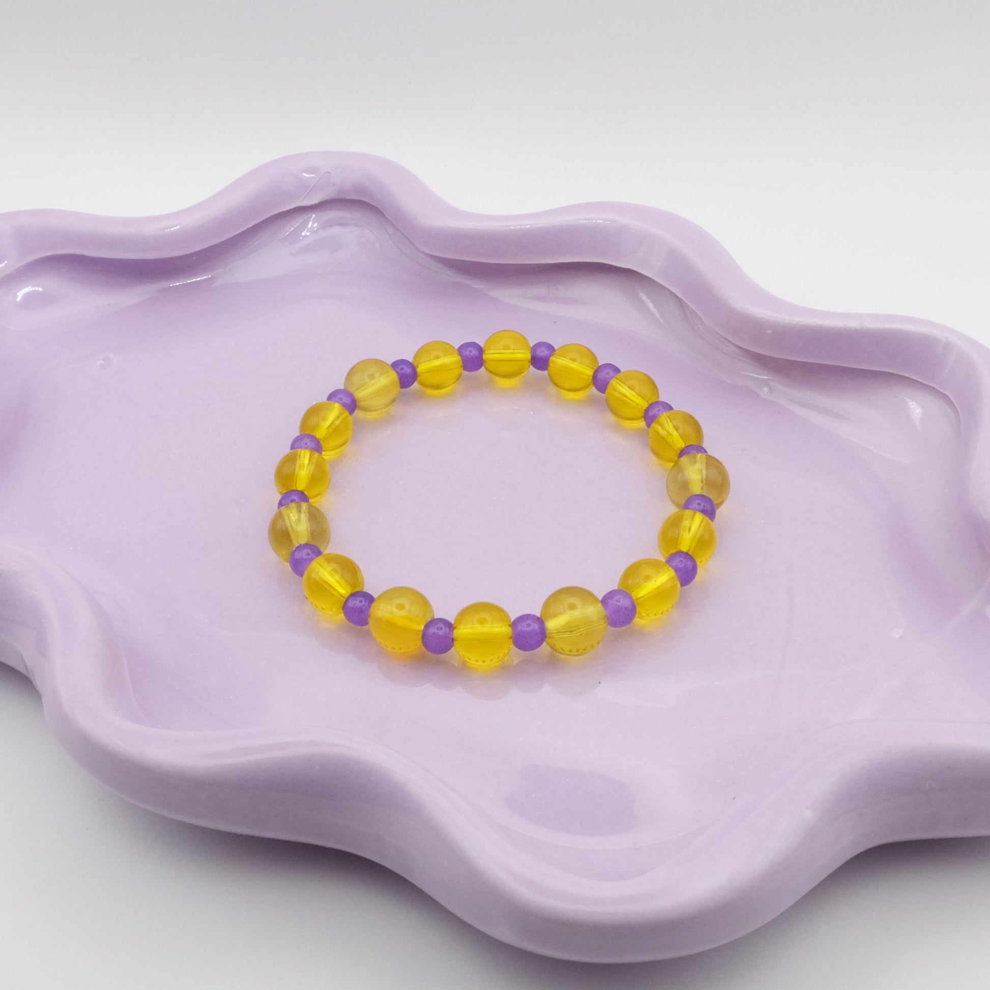 An elastic beaded bracelet featuring alternating 8mm transparent yellow glass beads and 6mm dark purple glass beads, displayed on a wavy lavender ceramic dish against a light backdrop.