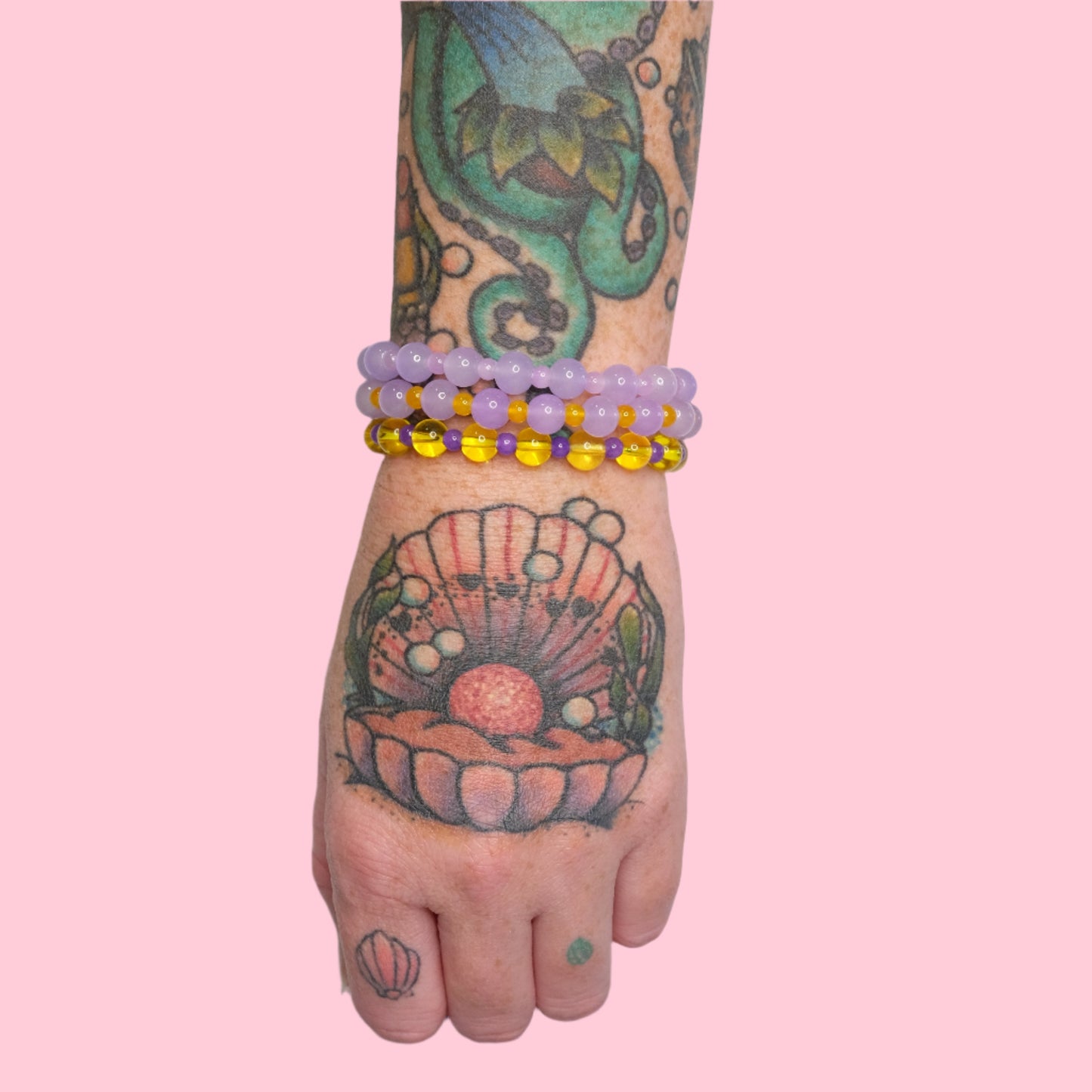 A tattooed arm with a vibrant octopus and seashell design is shown wearing multiple stacked elastic beaded bracelets in lavender and sunny yellow hues, set against a solid pink background.