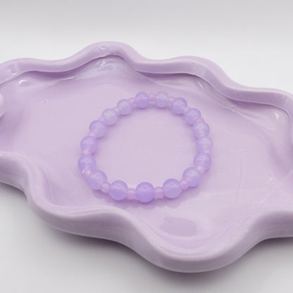 An elastic beaded bracelet composed of alternating 8mm lavender glass beads and 4mm lavender glass beads, resting on a wavy lavender ceramic dish in front of a neutral setting.