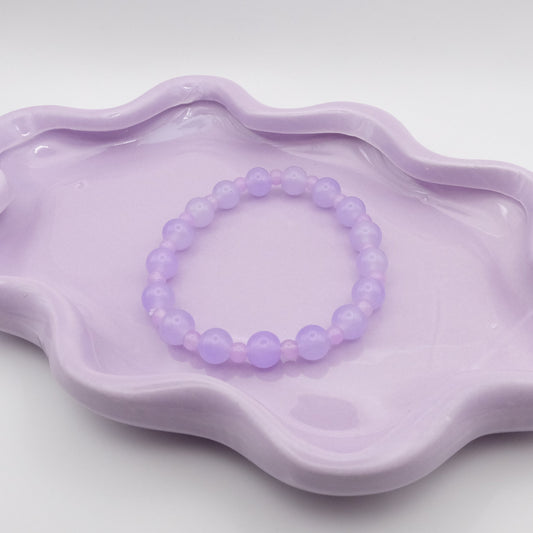 An elastic beaded bracelet composed of alternating 8mm lavender glass beads and 4mm lavender glass beads, resting on a wavy lavender ceramic dish in front of a neutral setting.