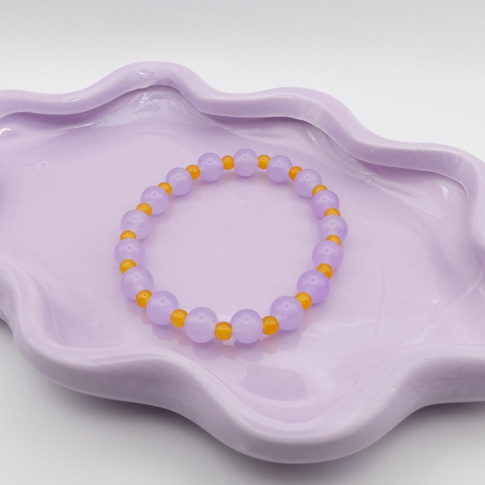 An elastic beaded bracelet showcasing alternating 8mm lavender purple glass beads and 4mm bright yellow glass beads, arranged on a wavy lavender ceramic dish with a clean background.