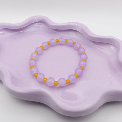 An elastic beaded bracelet showcasing alternating 8mm lavender purple glass beads and 4mm bright yellow glass beads, arranged on a wavy lavender ceramic dish with a clean background.