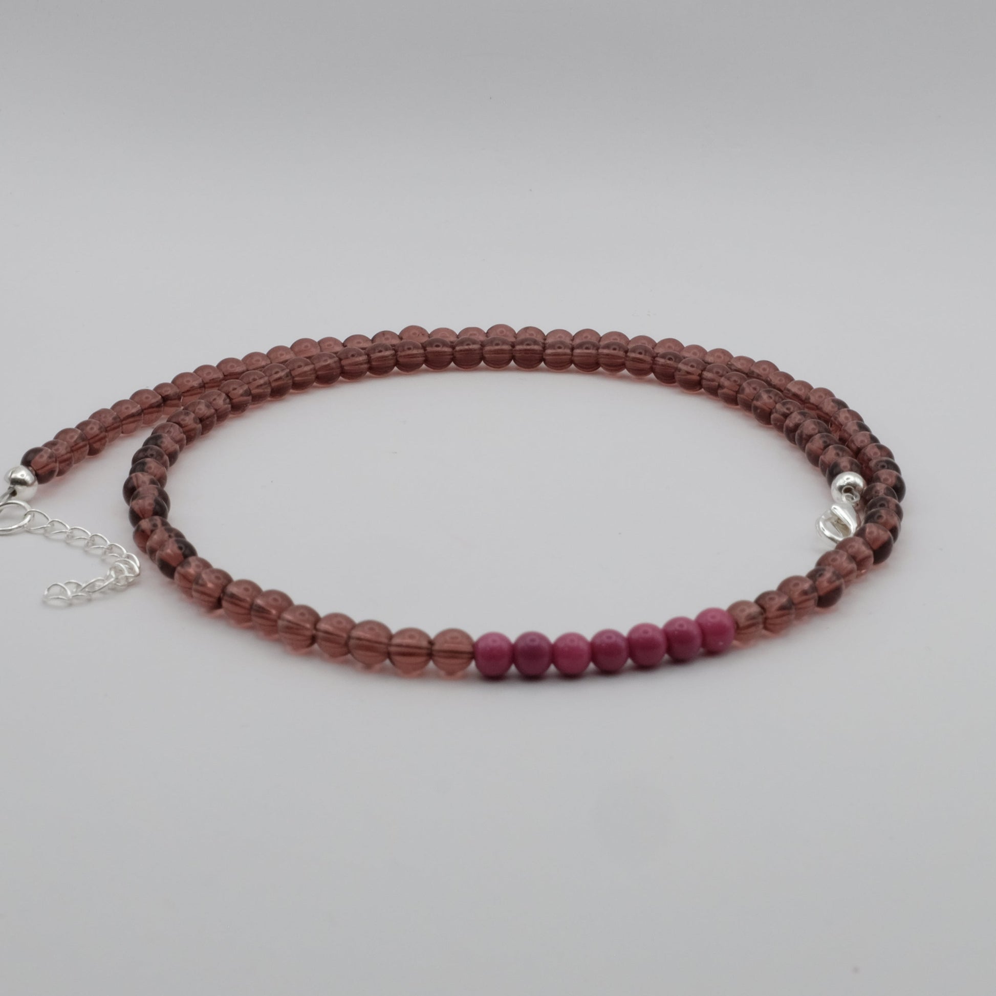 An elastic-style beaded necklace composed of smoky mauve translucent glass beads with a cluster of bright pink beads at the centre, displayed on a white marble jewellery stand.