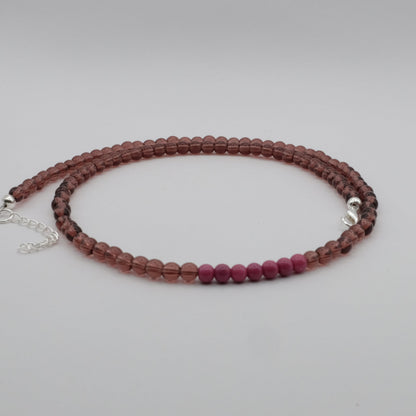 An elastic-style beaded necklace composed of smoky mauve translucent glass beads with a cluster of bright pink beads at the centre, displayed on a white marble jewellery stand.