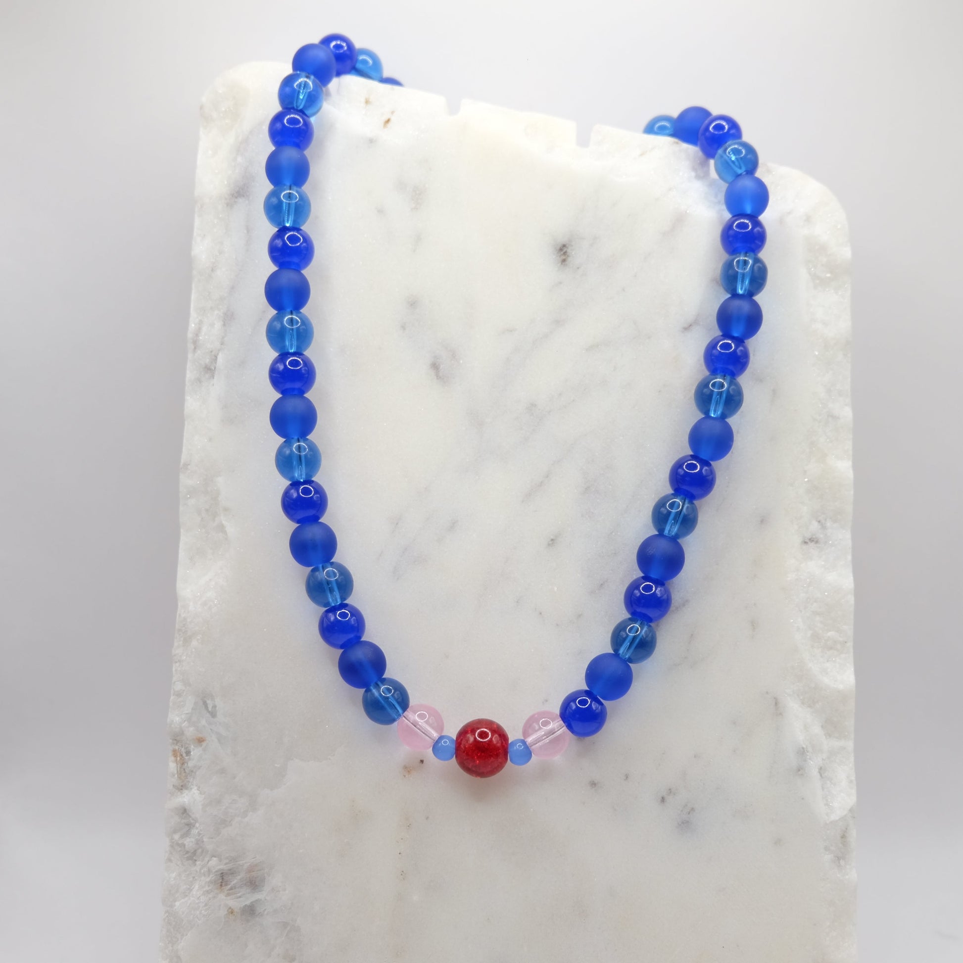 A beaded necklace composed of 8mm blue glass beads - featuring a red glitter centre bead with pink and blue accents, shown on a white marble background.