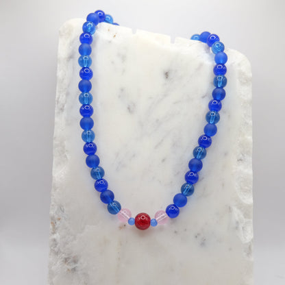 A beaded necklace composed of 8mm blue glass beads - featuring a red glitter centre bead with pink and blue accents, shown on a white marble background.