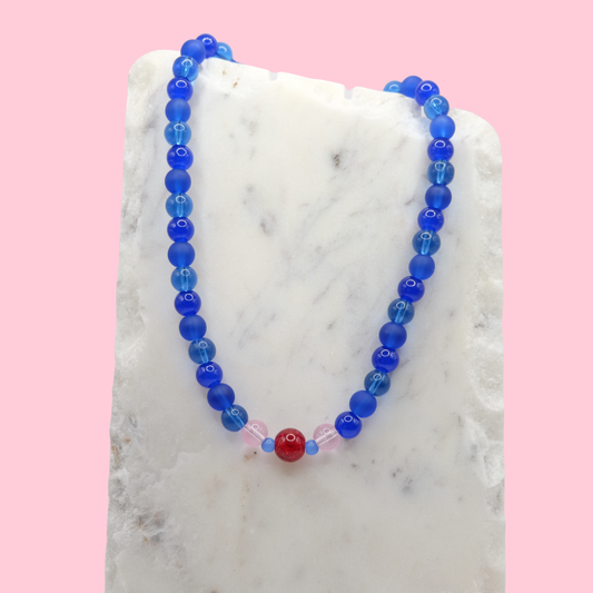 Blue beaded necklace with a red accent bead on a marble surface with a pink background.