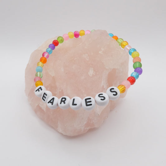An elastic beaded bracelet composed of colourful 4mm glass beads in red,  orange, yellow, green, aqua, blue,  purple and pink with white acrylic letter beads spelling “FEARLESS,” displayed on a pale pink crystal rock.