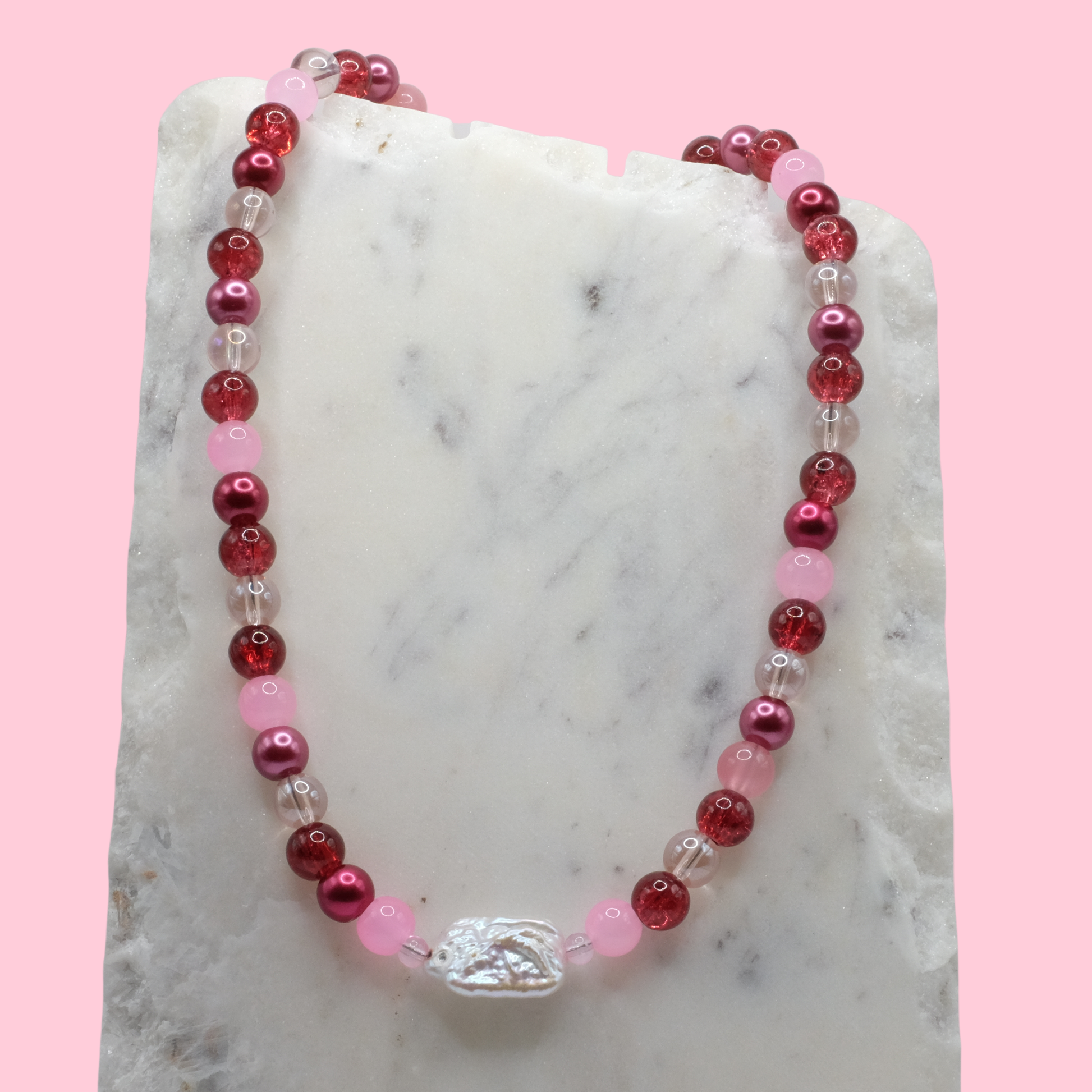 Beaded necklace with red, pink, and clear beads on a marble surface with a pink background