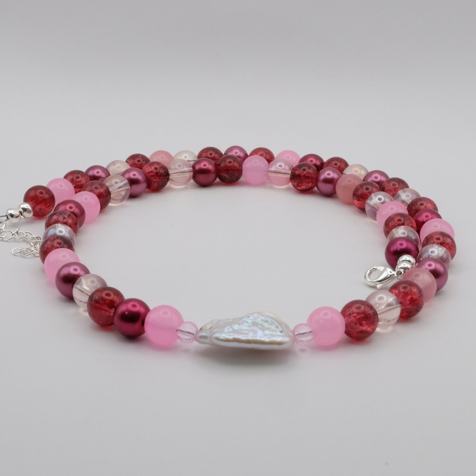 A handmade beaded necklace featuring pink, berry and clear glass beads with a freshwater pearl centrepiece, displayed on a white marble jewellery stand.