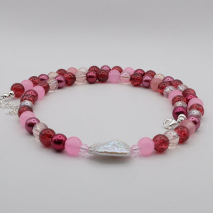 A handmade beaded necklace featuring pink, berry and clear glass beads with a freshwater pearl centrepiece, displayed on a white marble jewellery stand.