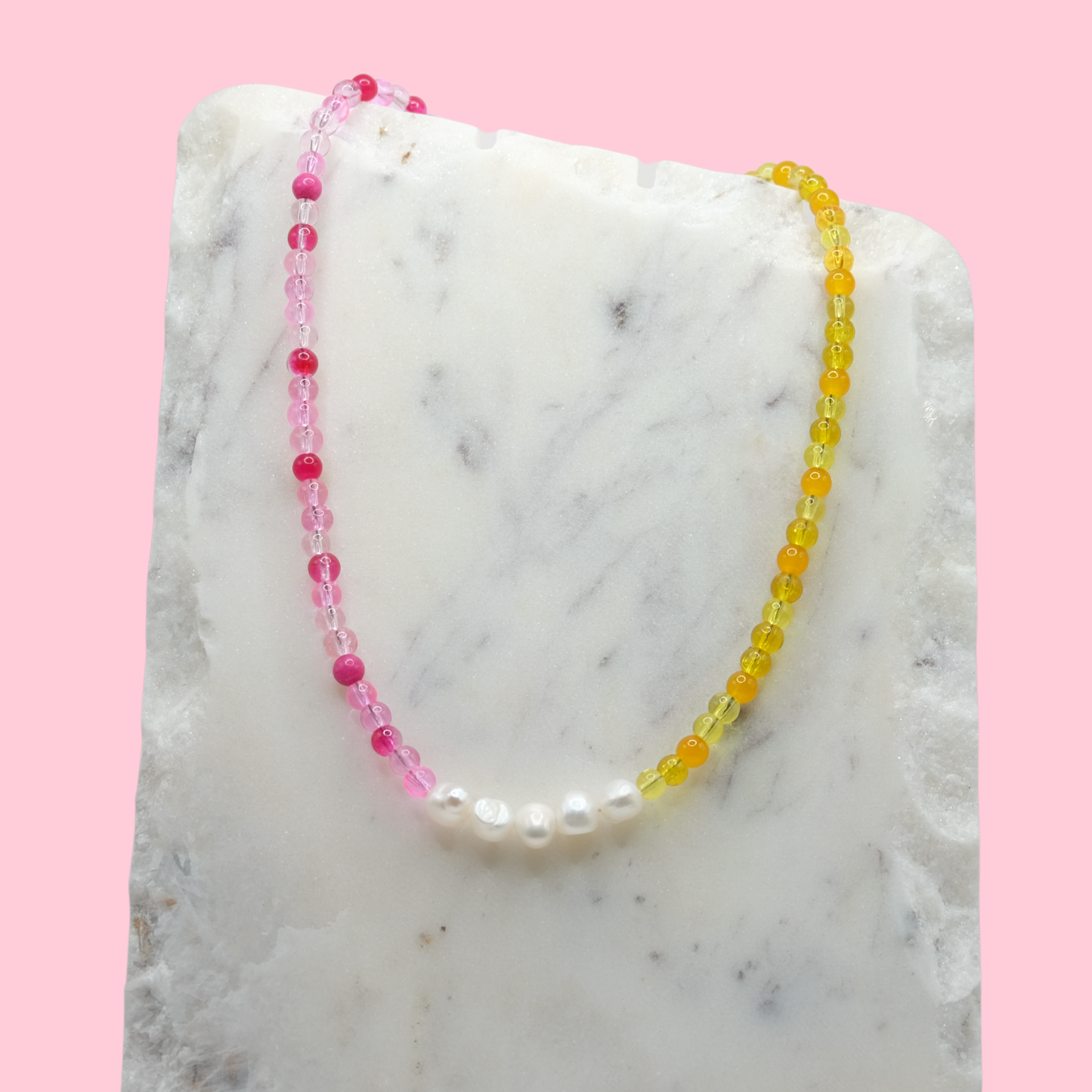 A handmade beaded necklace composed of 4mm pink and yellow glass beads with five freshwater pearl nuggets at the centre, displayed on a white marble slab against a pink background.