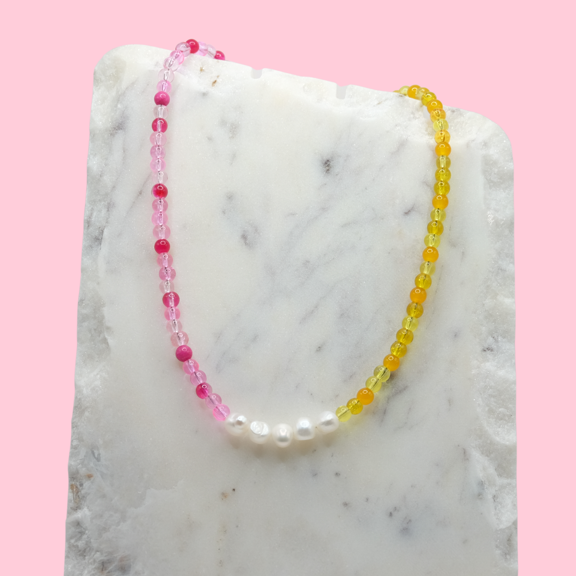 A handmade beaded necklace composed of 4mm pink and yellow glass beads with five freshwater pearl nuggets at the centre, displayed on a white marble slab against a pink background.