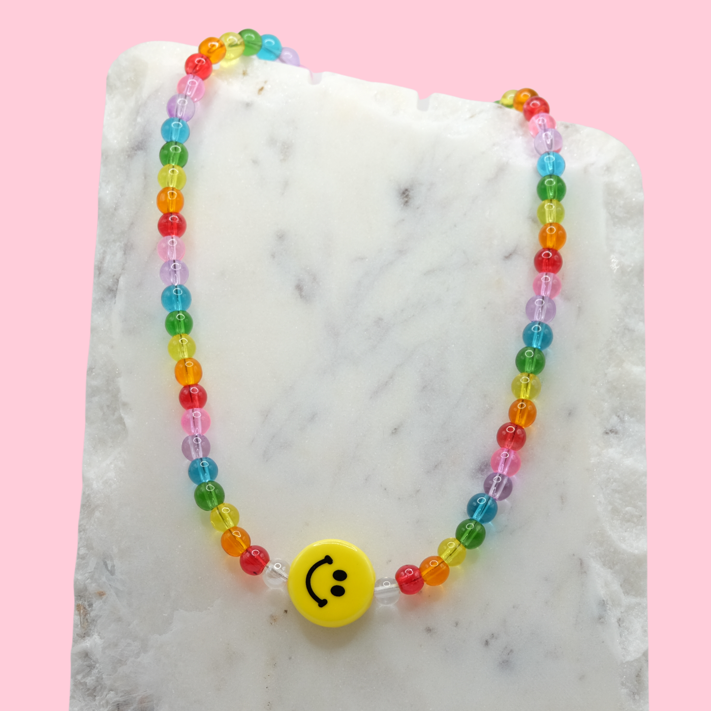 A handmade beaded necklace composed of translucent 6mm glass beads in rainbow colours, featuring a yellow ceramic smiley face bead at the centre, displayed on a white marble slab.