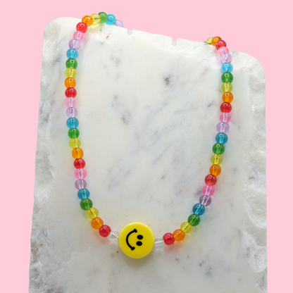 A handmade beaded necklace composed of translucent 6mm glass beads in rainbow colours, featuring a yellow ceramic smiley face bead at the centre, displayed on a white marble slab.