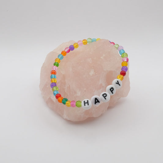 An elastic beaded bracelet composed of multicoloured 4mm glass beads with white acrylic letter beads spelling “HAPPY”, displayed on a pale pink crystal rock.