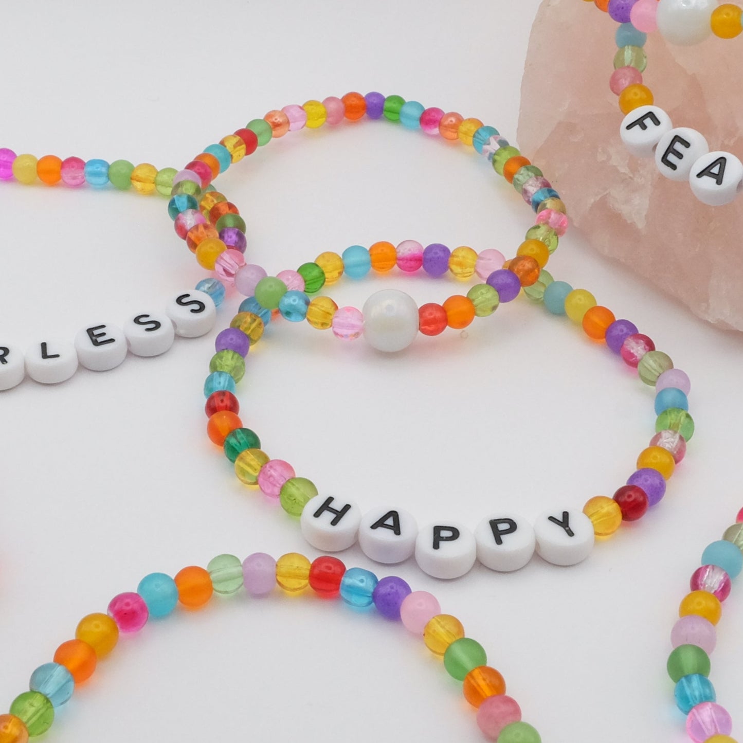A flat lay of  multiple elastic beaded bracelet composed of multicoloured 4mm glass beads with white acrylic letter beads spelling “HAPPY,” displayed on a pale pink crystal rock.